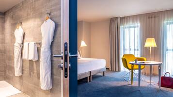 A hotel room with a bed, a chair, a table, a lamp, and a red handbag.