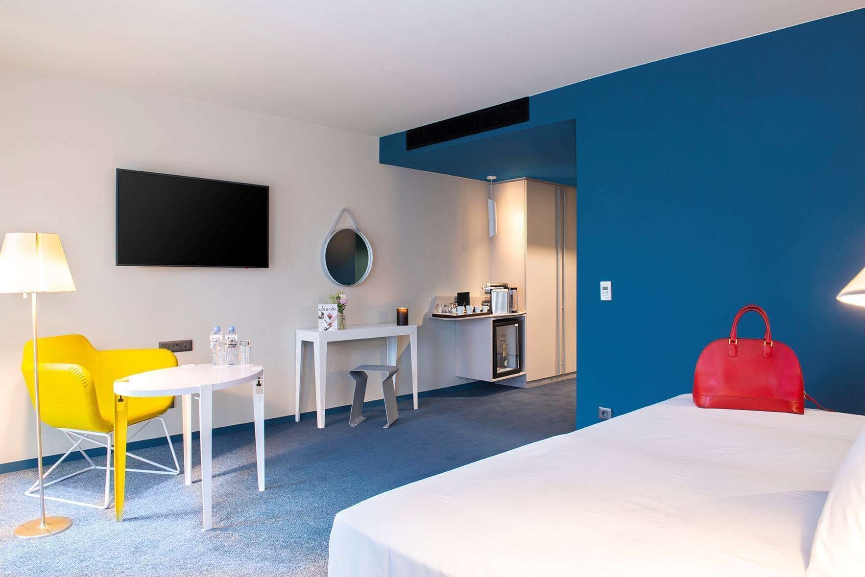 Une chambre d’hôtel avec un lit, un bureau, une chaise, une télévision, un miroir et un sac à main rouge.
