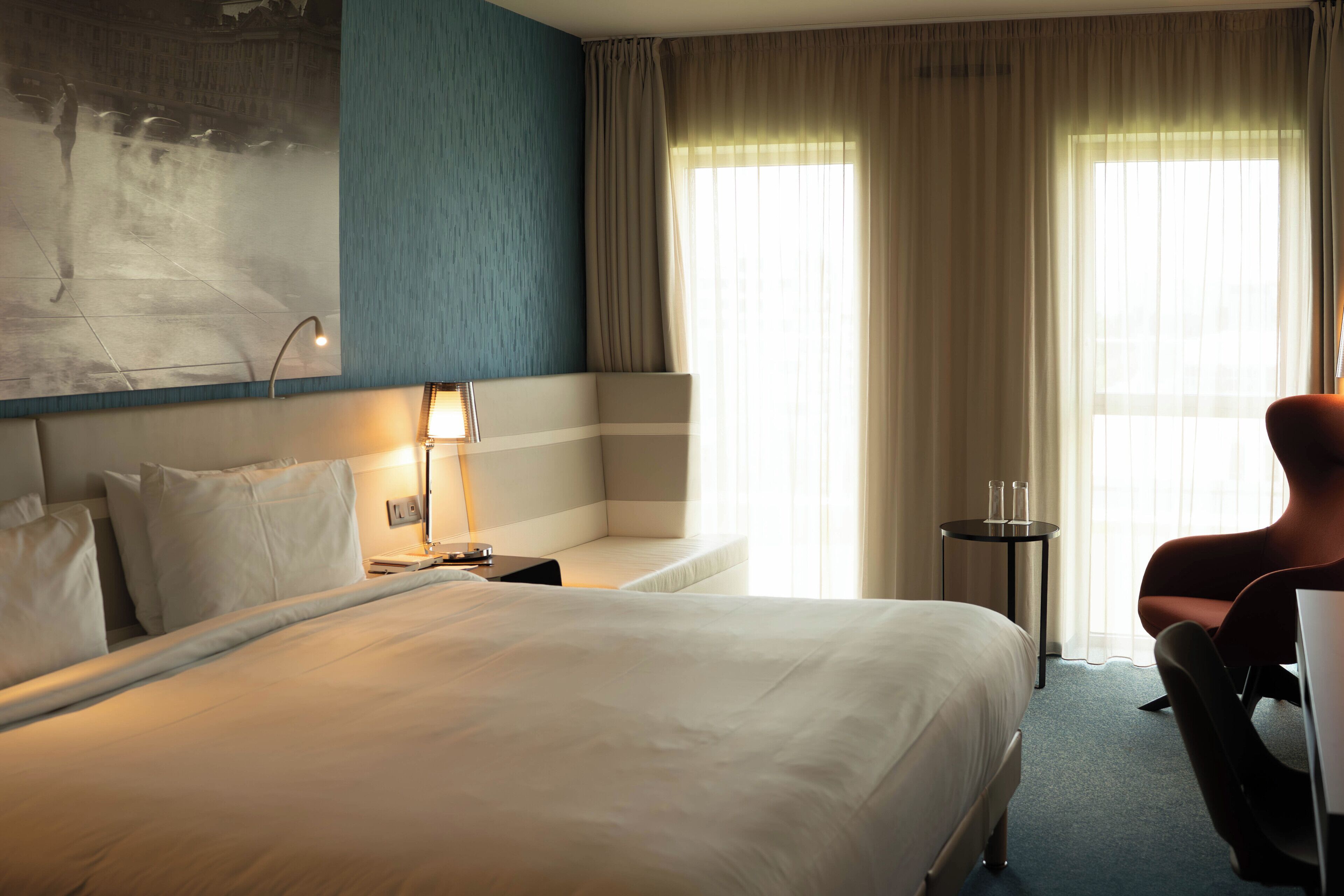 A hotel room with a large bed, a bedside table with a lamp, a chair, and a view of the city through the window.