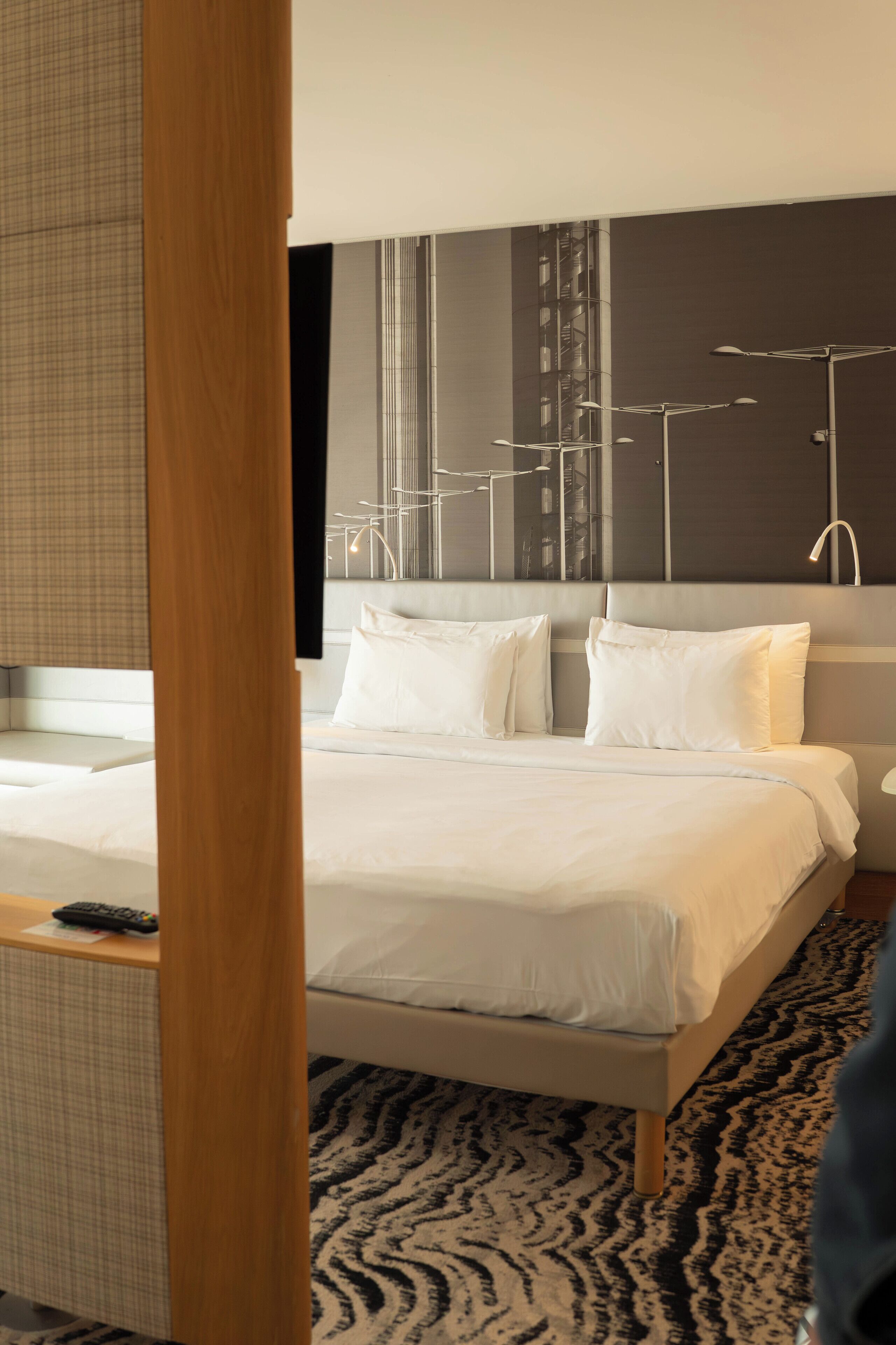 Una habitación de hotel moderna con una cama grande, un cabecero con lámpara, una mesita de noche y una alfombra estampada.