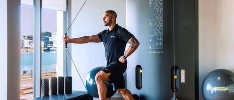 Un homme fait de l'exercice sur une plateforme en marchant, en utilisant un appareil à câbles, un sac de frappe et un grand ballon d'exercice dans une salle de sport offrant une vue sur la ville.