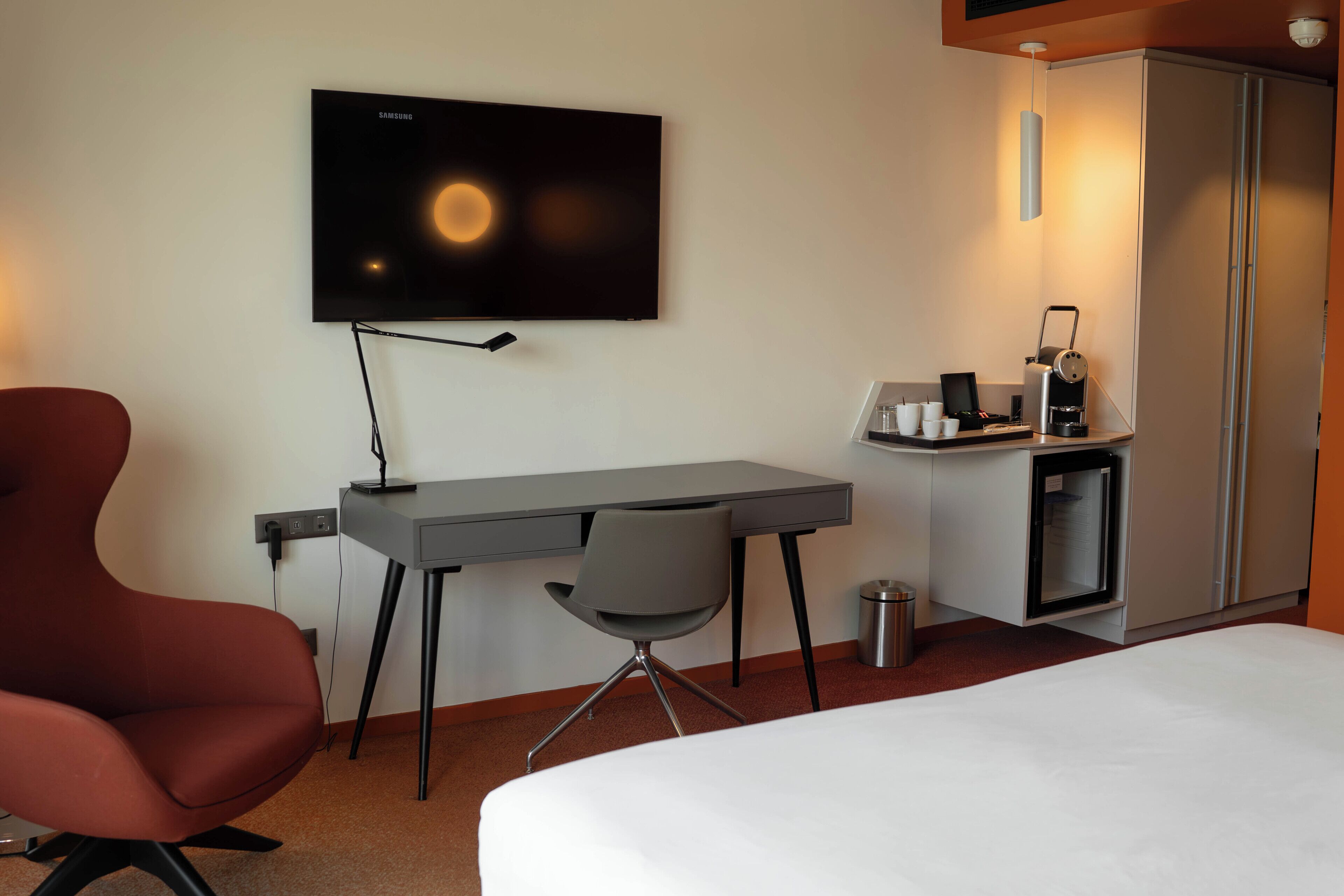 Habitación de hotel con cama, escritorio, silla, televisor, refrigerador pequeño y cafetera.