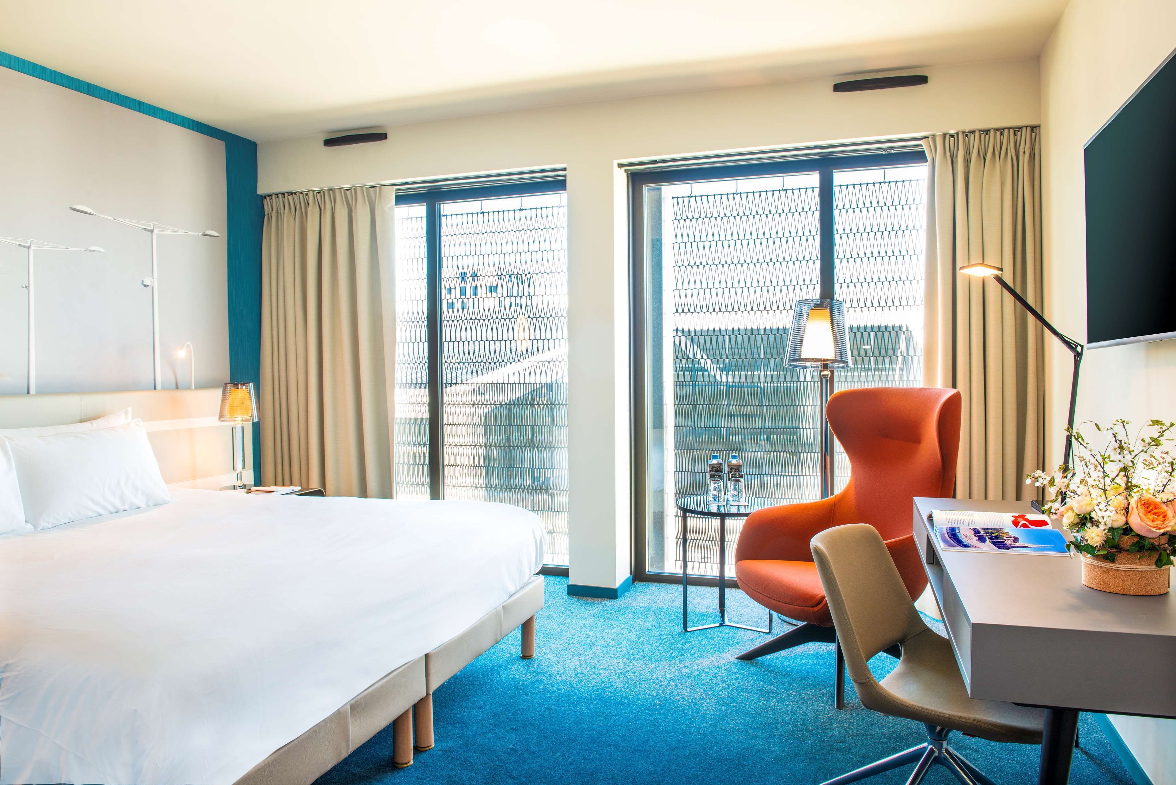 Une chambre d’hôtel avec un grand lit, un bureau avec une chaise, une lampe et une vue sur la ville grâce à une grande fenêtre.