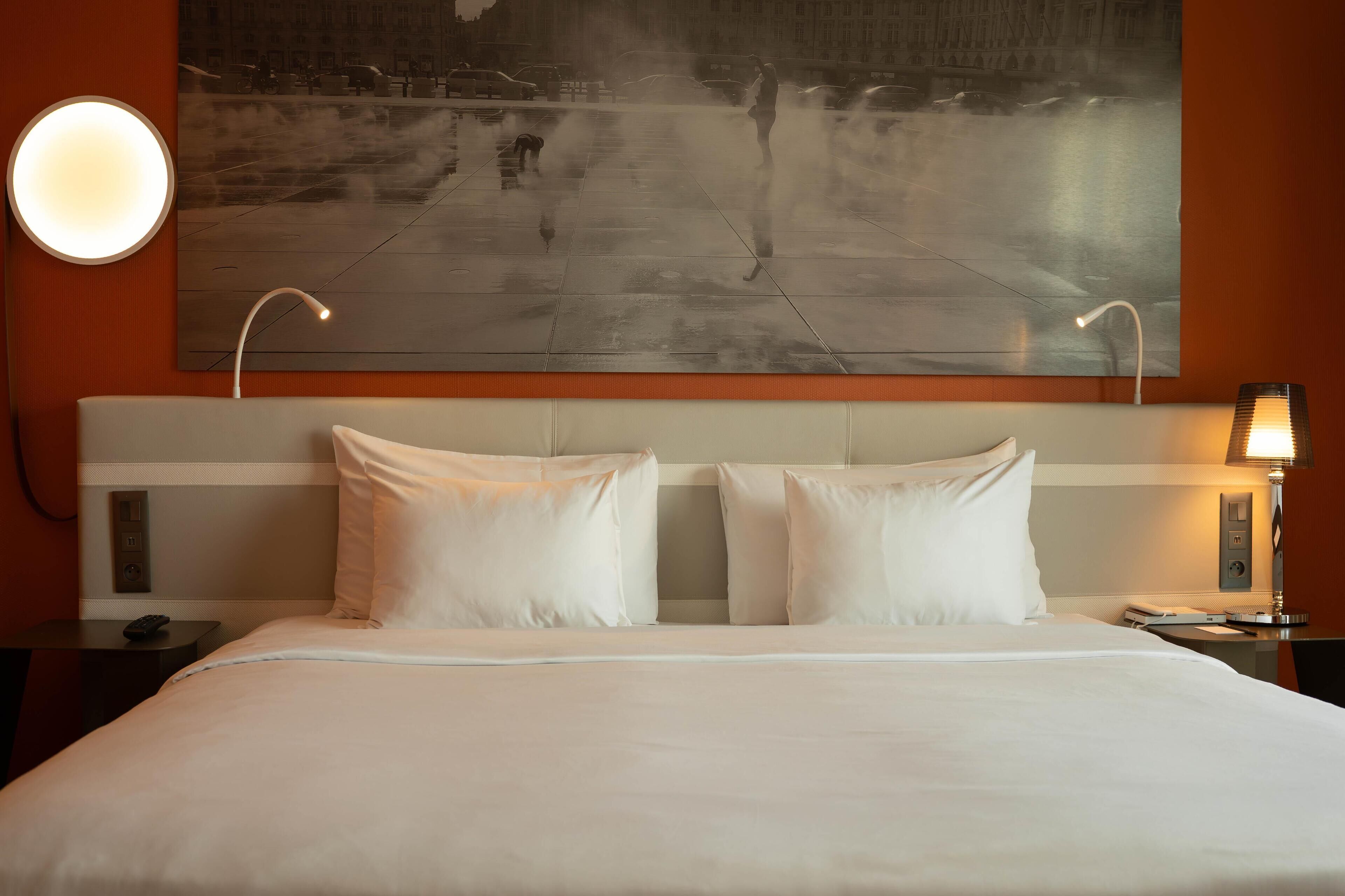 Habitación de hotel con una cama grande, dos lámparas de noche, una mesita de noche y una obra de arte en la pared que representa un paisaje urbano.