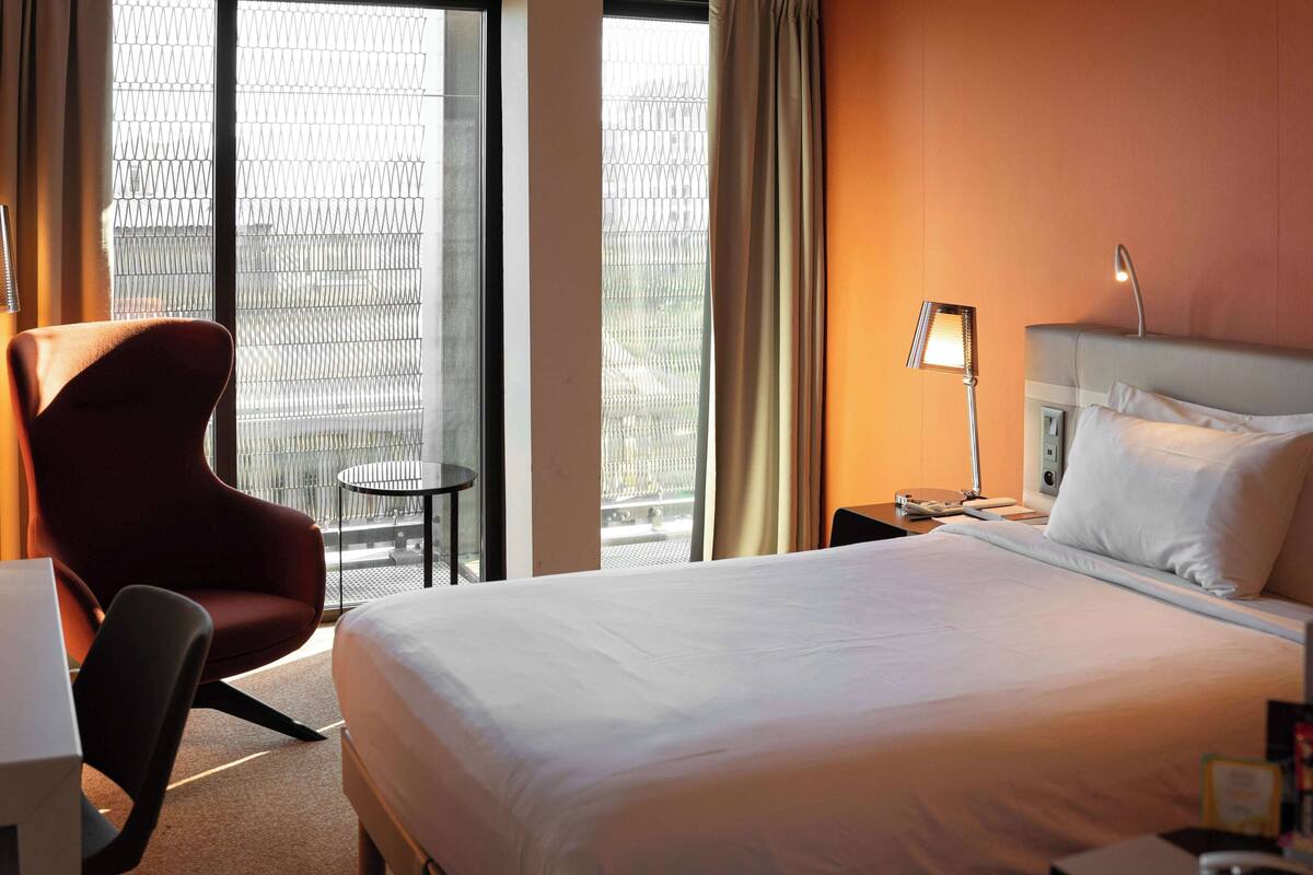 Une chambre d’hôtel avec un grand lit, un bureau, une chaise, une lampe et une fenêtre donnant sur l’extérieur, protégée par un moustiquaire métallique.