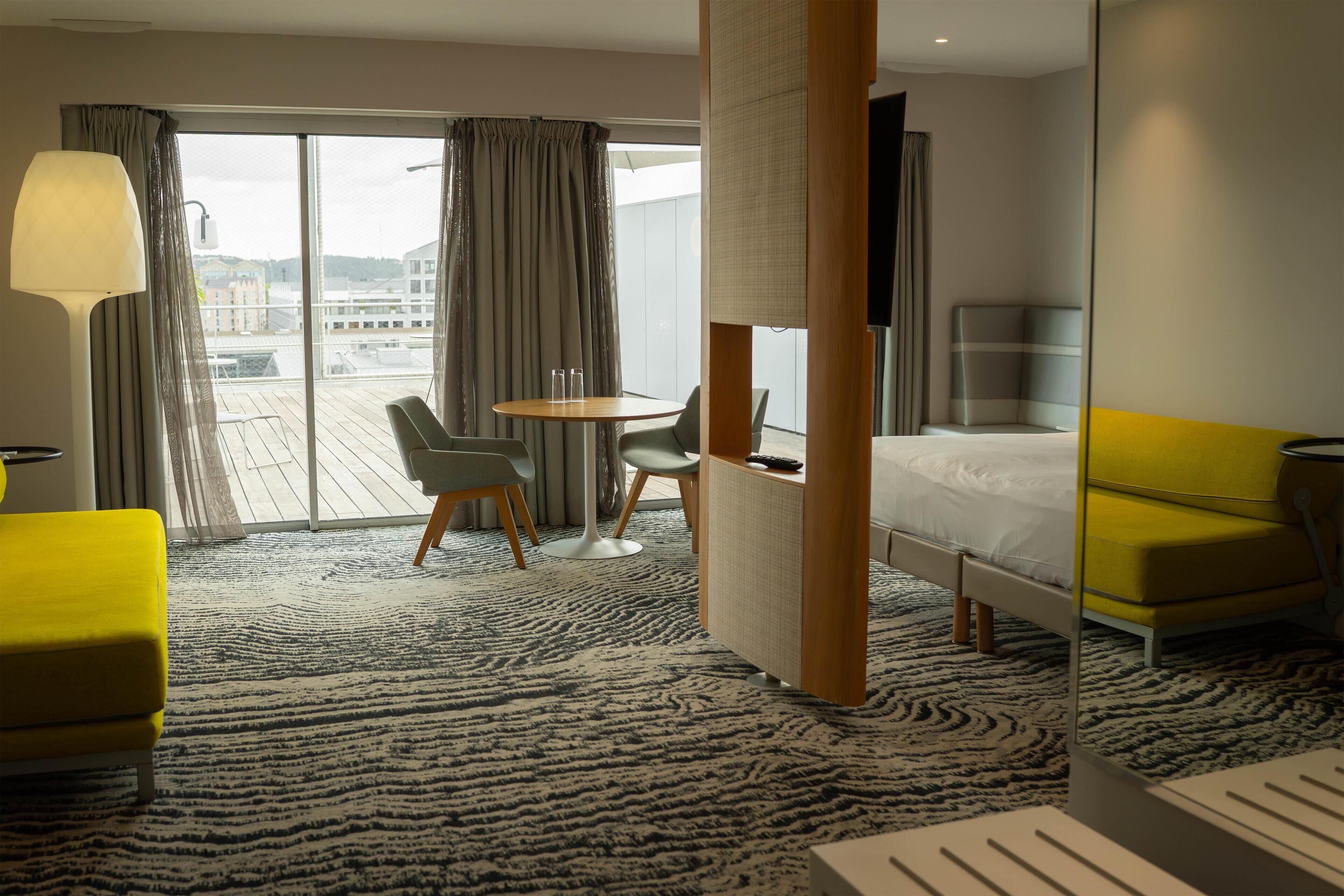 Ein Hotelzimmer mit einem Bett, einem gelben Sofa, einem runden Tisch mit Stühlen, einem Schreibtisch und einem großen Fenster mit Vorhängen.
