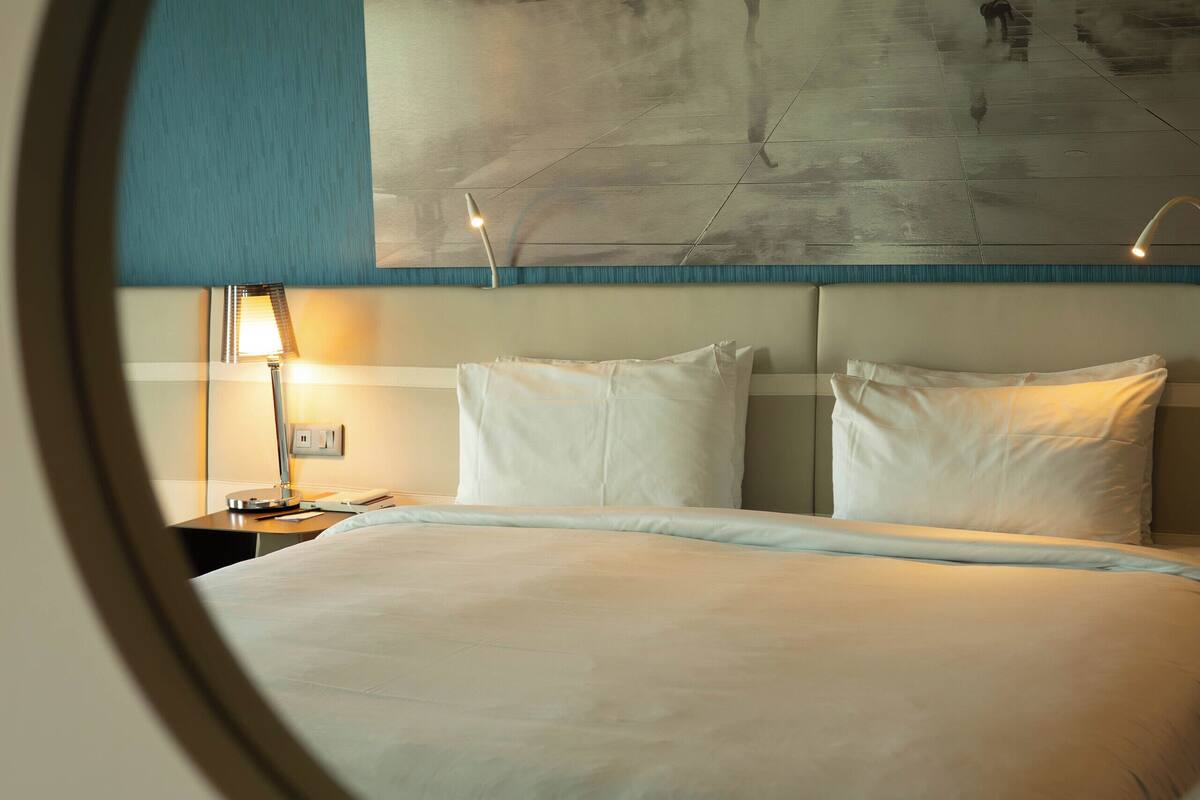 Une chambre d’hôtel avec un lit, une table de chevet, une lampe et une grande affiche accrochée au mur.