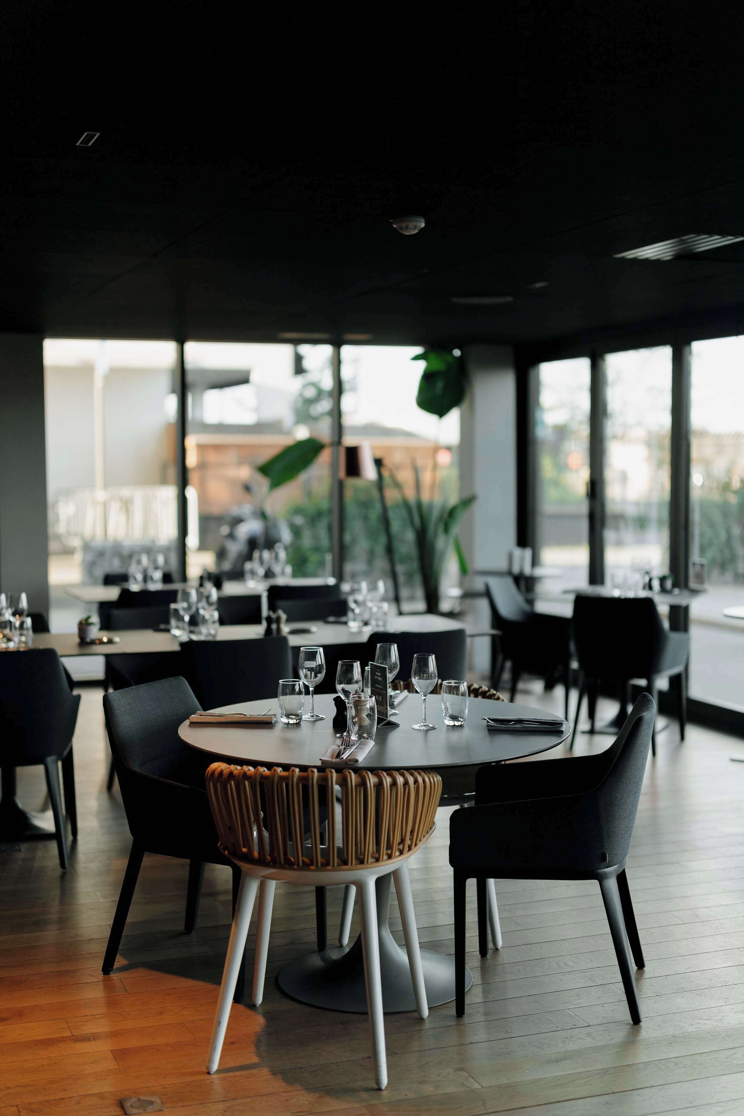 Een restaurant met ronde tafels, zwarte stoelen en uitzicht op de straat door grote ramen.