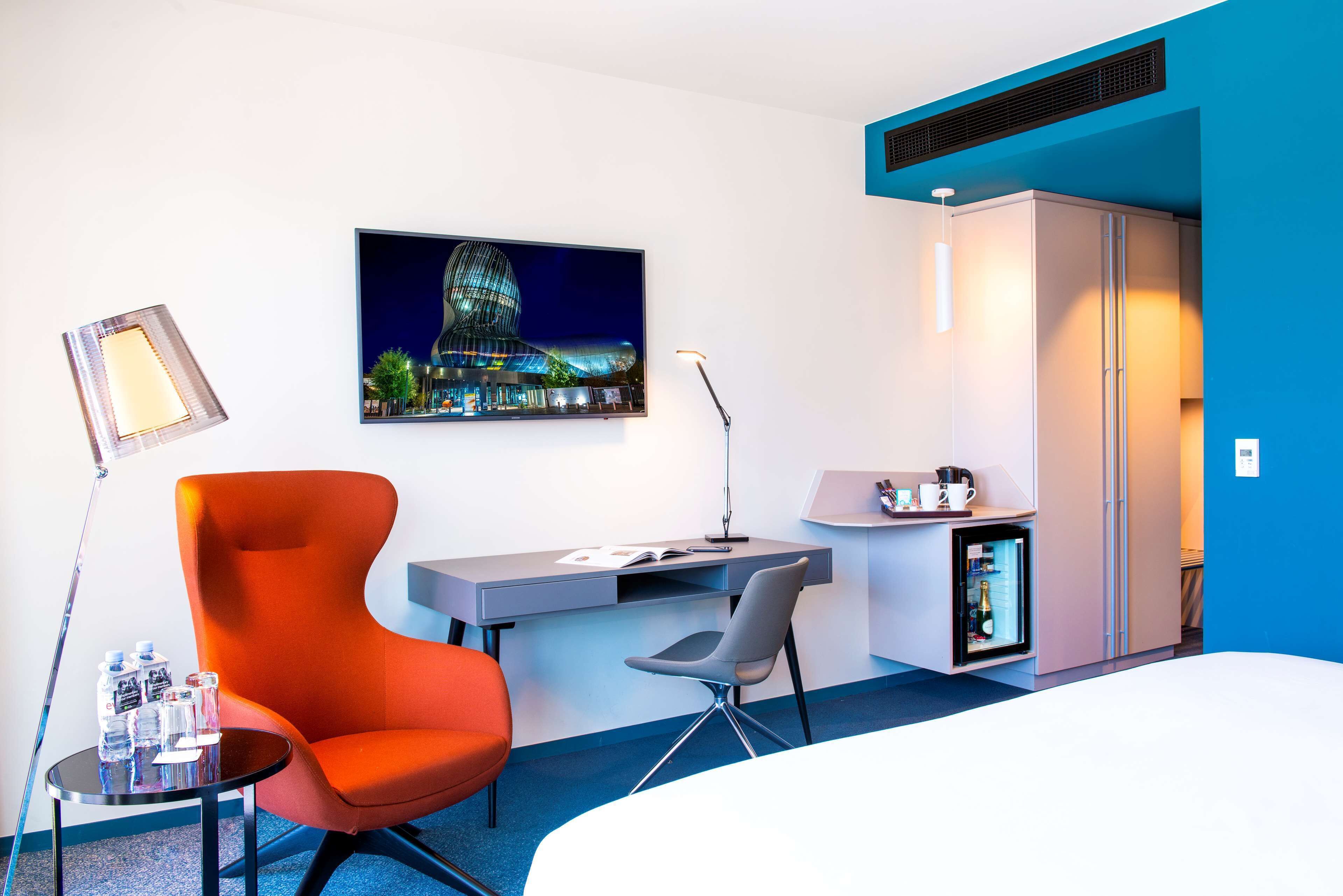 A hotel room with a desk, a chair, a bed, a refrigerator, a mini-fridge, a lamp, and a picture on the wall.