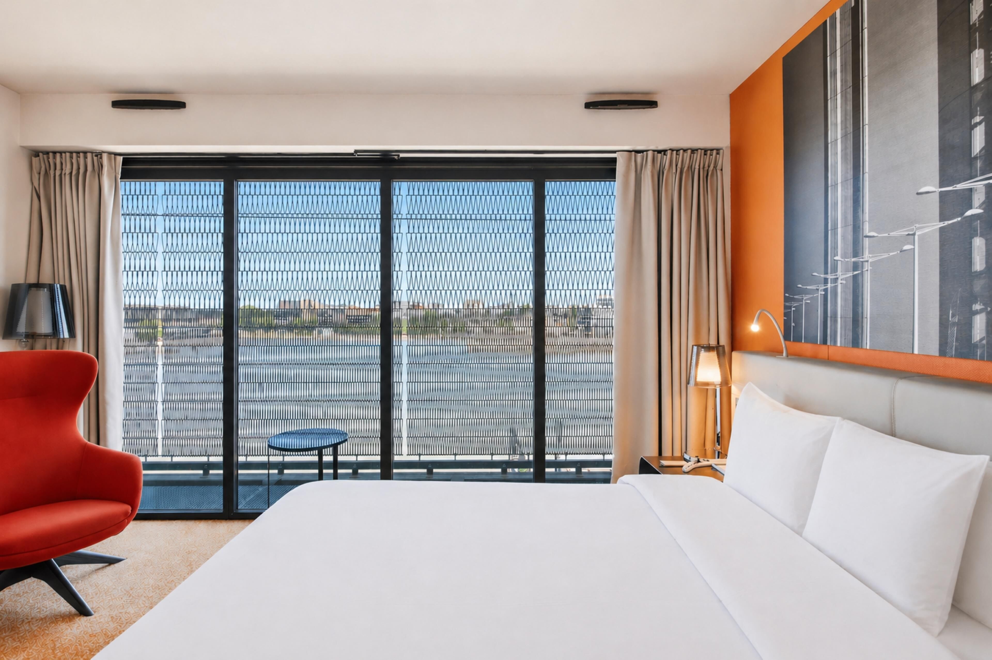 Habitación de hotel con una cama grande, una silla roja, una mesita y vistas a través de un ventanal amplio.