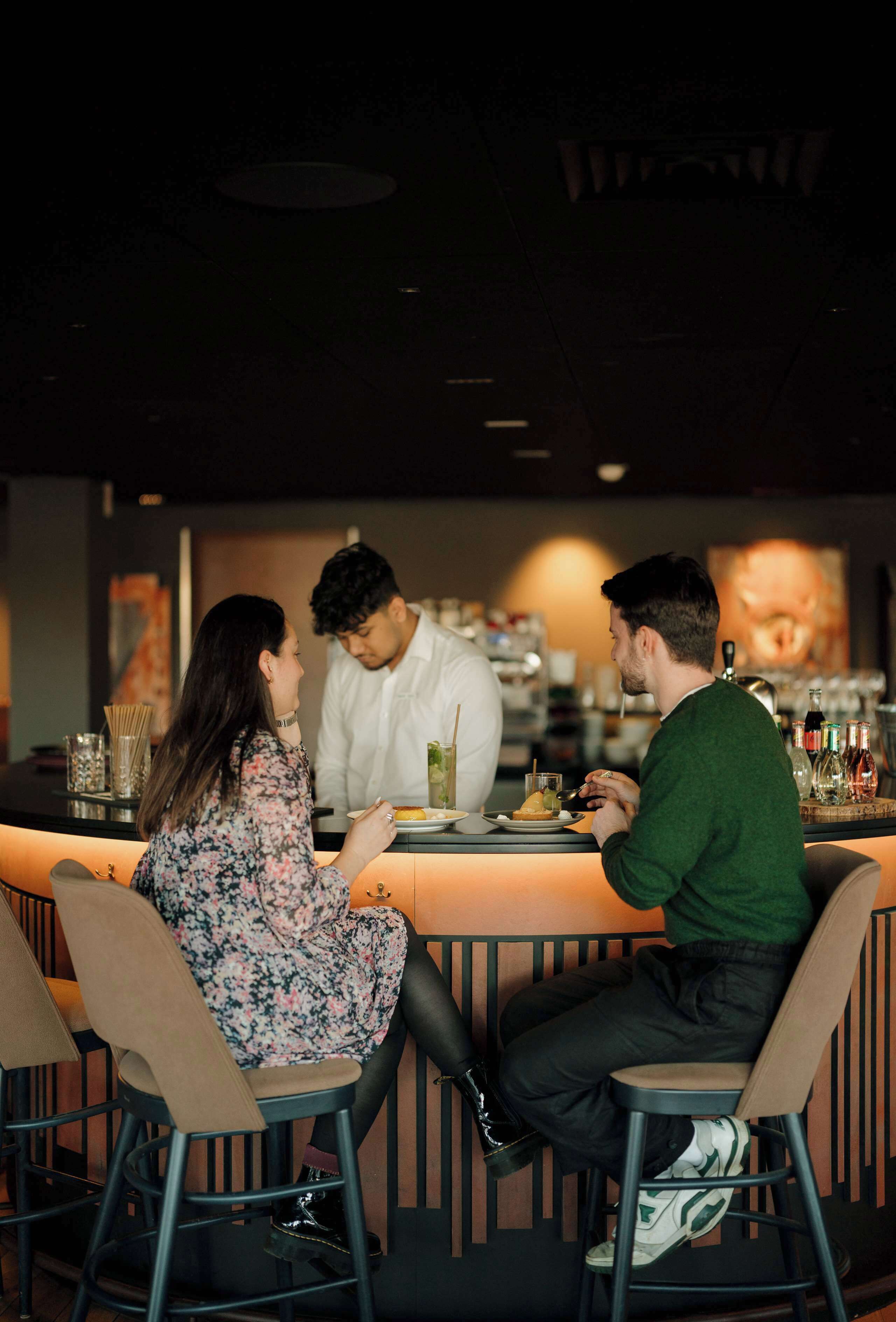 A bar setting with a counter, stools, and a person serving drinks.