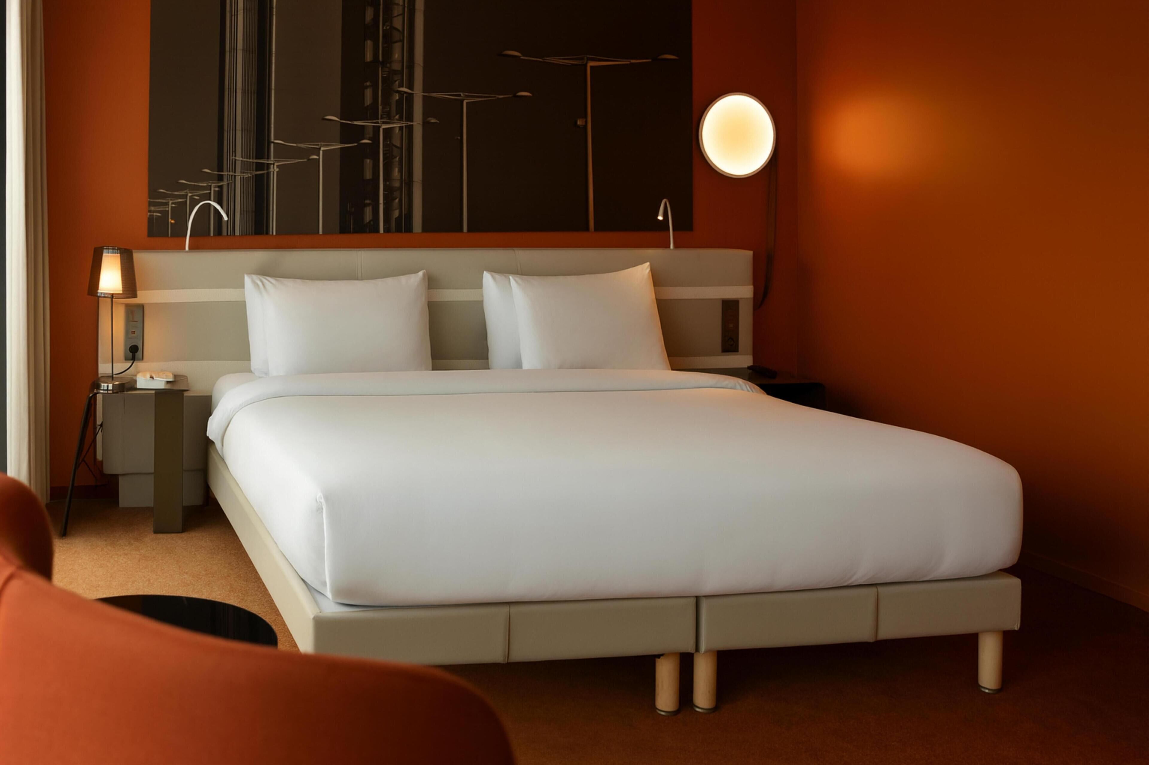 A modern hotel room with a large bed, a nightstand, a lamp, and a wall-mounted light.