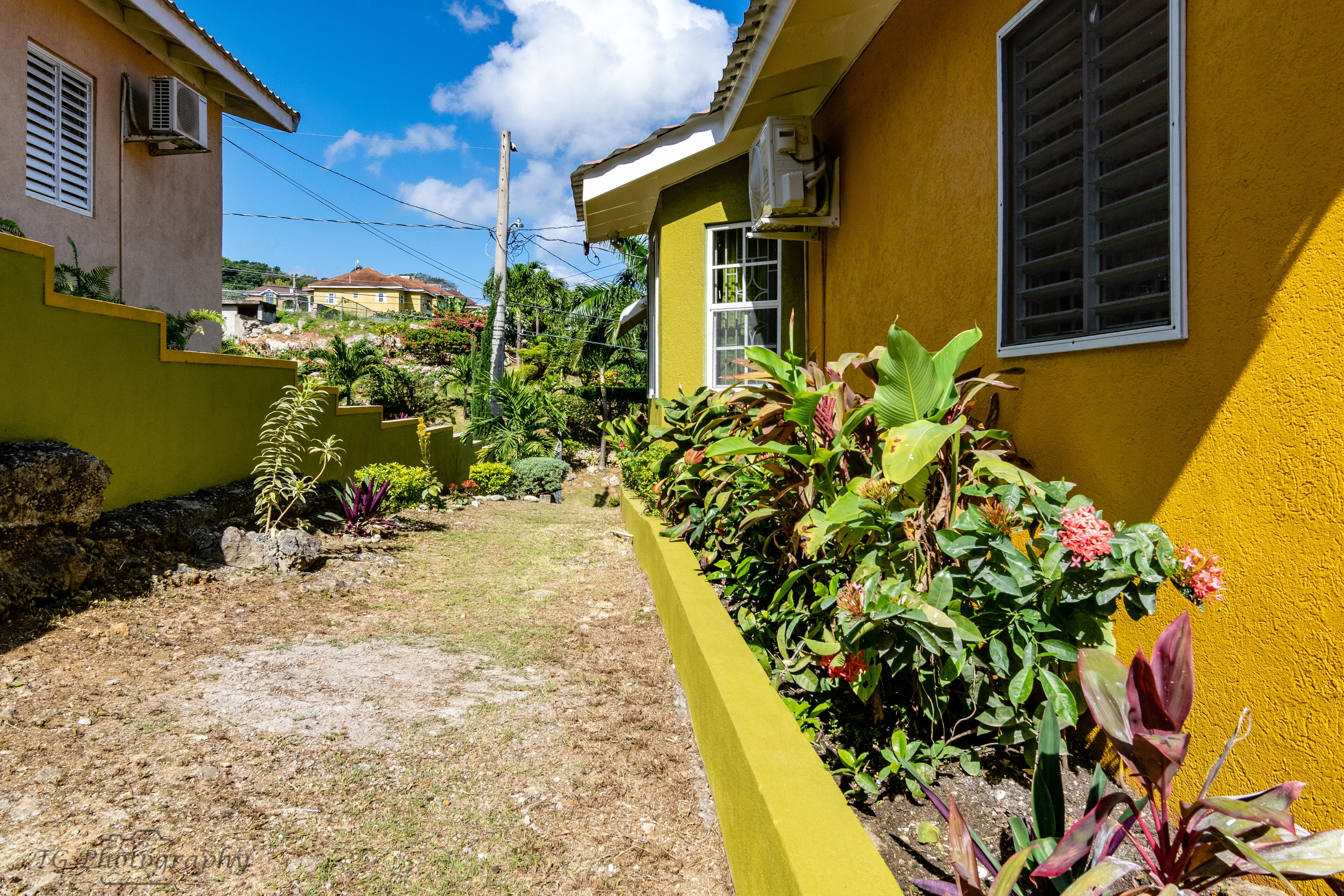 A yellow building with a small garden area, a paved path, and a stone wall.