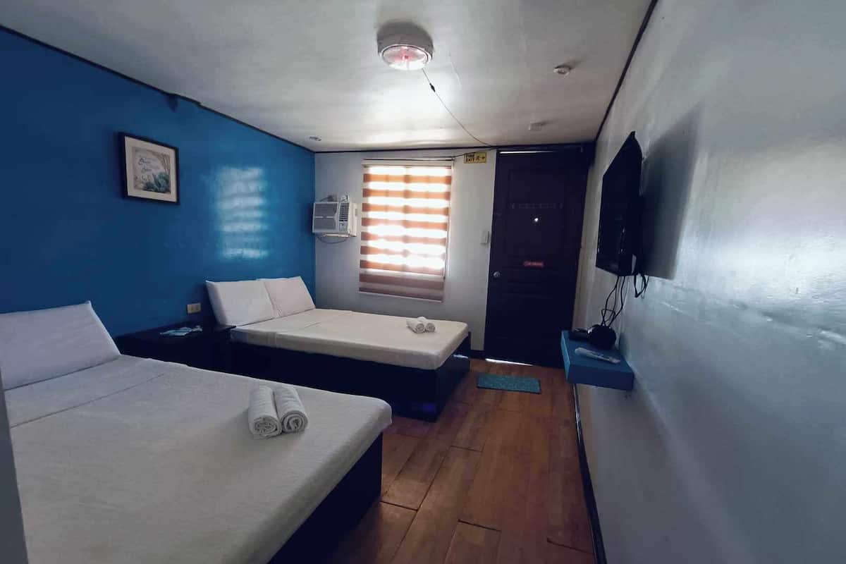 A small, compact room with two single beds, a ceiling fan, a mounted TV, and a window with blinds.