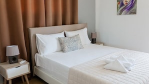 A neatly made bed with white linens and a quilted blanket, a bedside table with a lamp, and a painting on the wall. - Steliana 2 (Malevizi)
