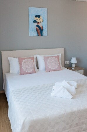 A neatly made bed with white bedding and pink decorative pillows, a bedside table with a lamp, and a painting on the wall.