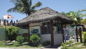 A small building with a thatched roof, labeled 'Clube', surrounded by greenery and palm trees. - Apto Cabo Frio Marina Clube (Sao Pedro da Aldeia)