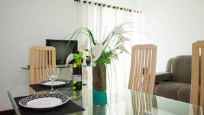 A dining area with a glass table, chairs, a vase with flowers, and a television in the background. - Apto Cabo Frio Marina Clube (Sao Pedro da Aldeia)
