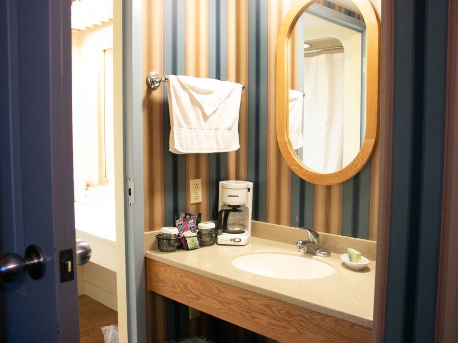 Un bagno d'albergo con un mobiletto vanità in legno, uno specchio rotondo, una macchina per caffè e un asciugamano appeso al muro.