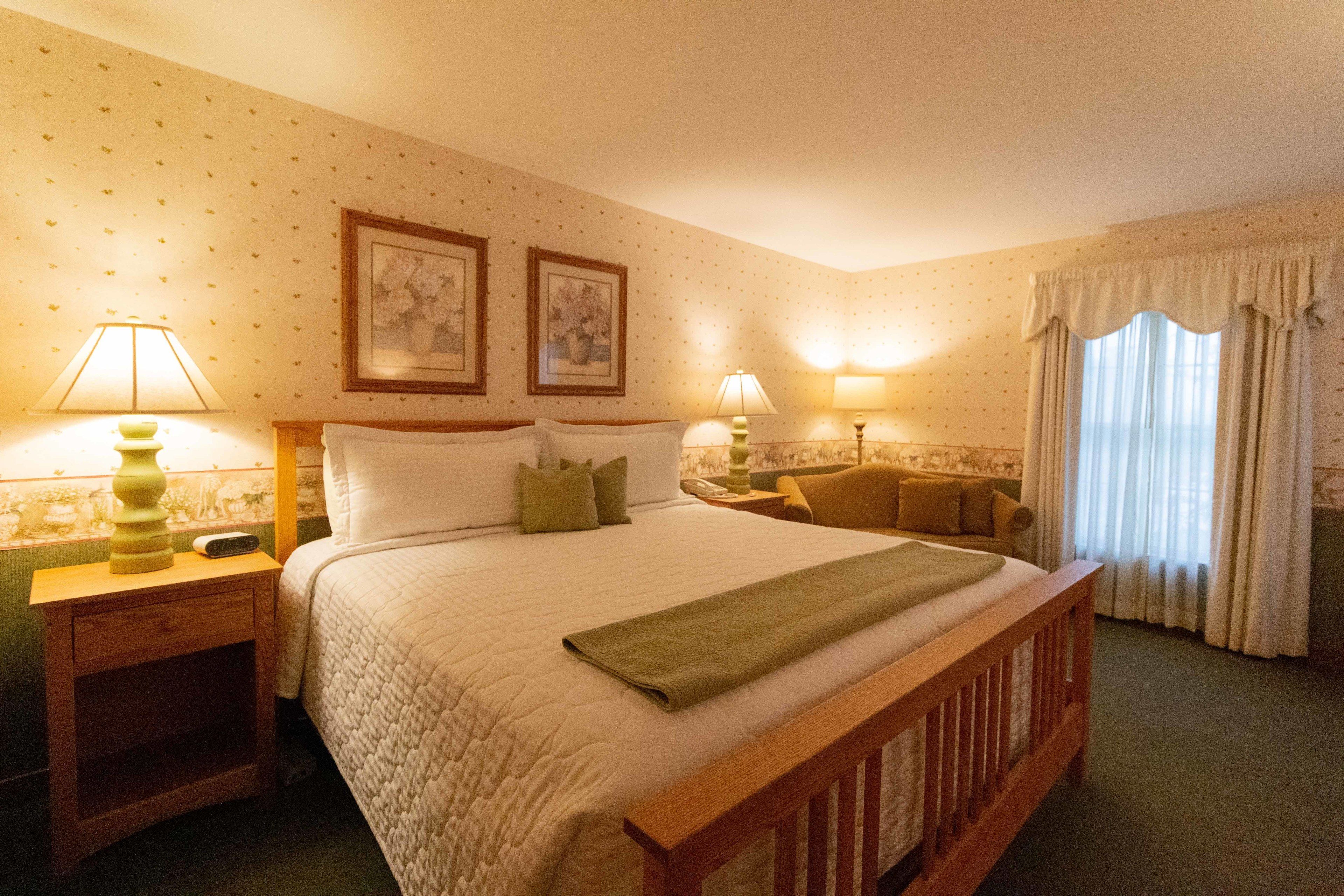 Habitación de hotel con una cama grande, dos mesitas de noche con lámparas, una silla y un ventanal con cortinas.
