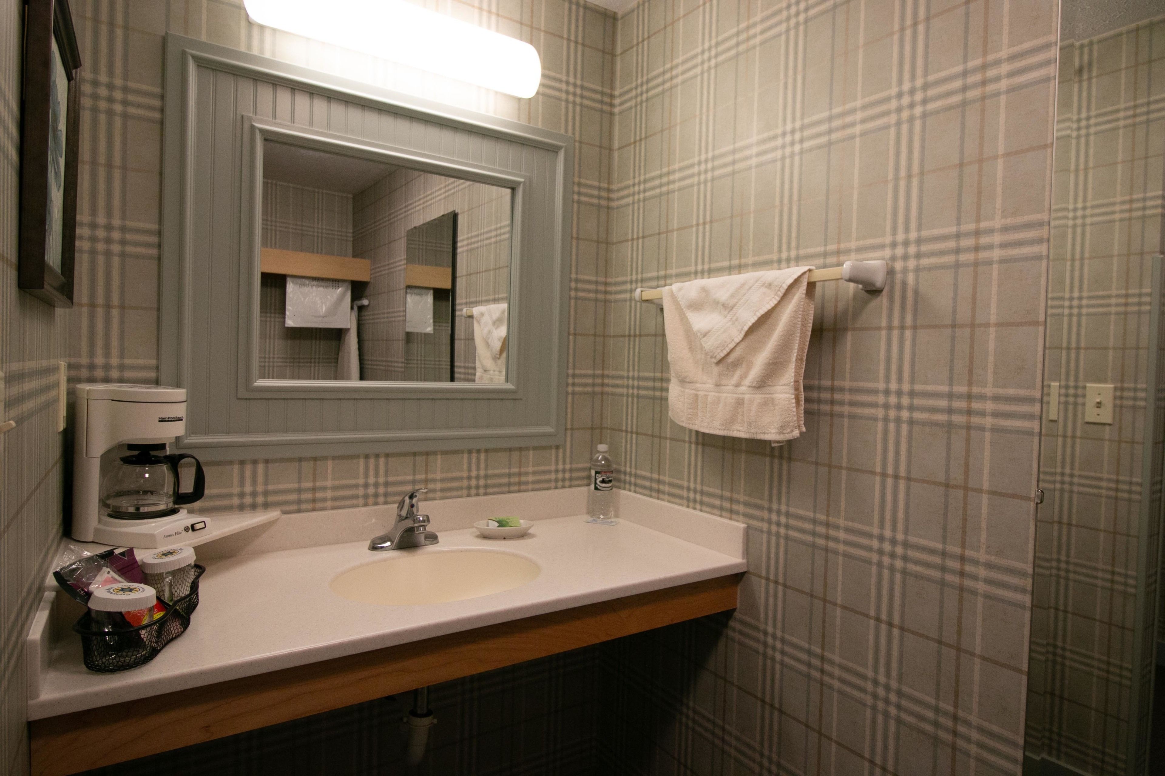 Un bagno con lavandino bianco, specchio, porta asciugamani e macchina per il caffè.