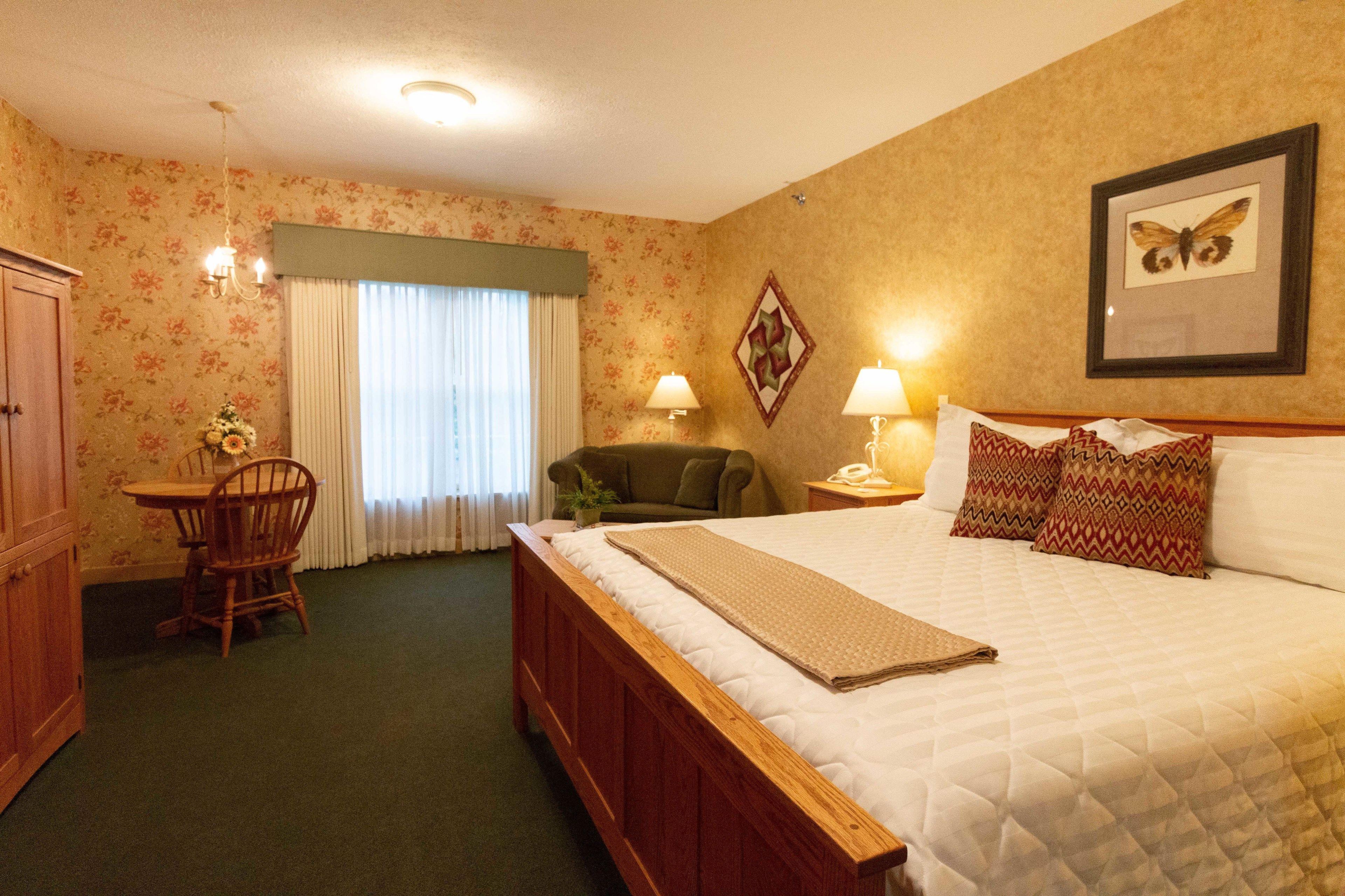 A hotel room with a large bed, a desk with a chair, a small table with a plant, and a window with curtains.