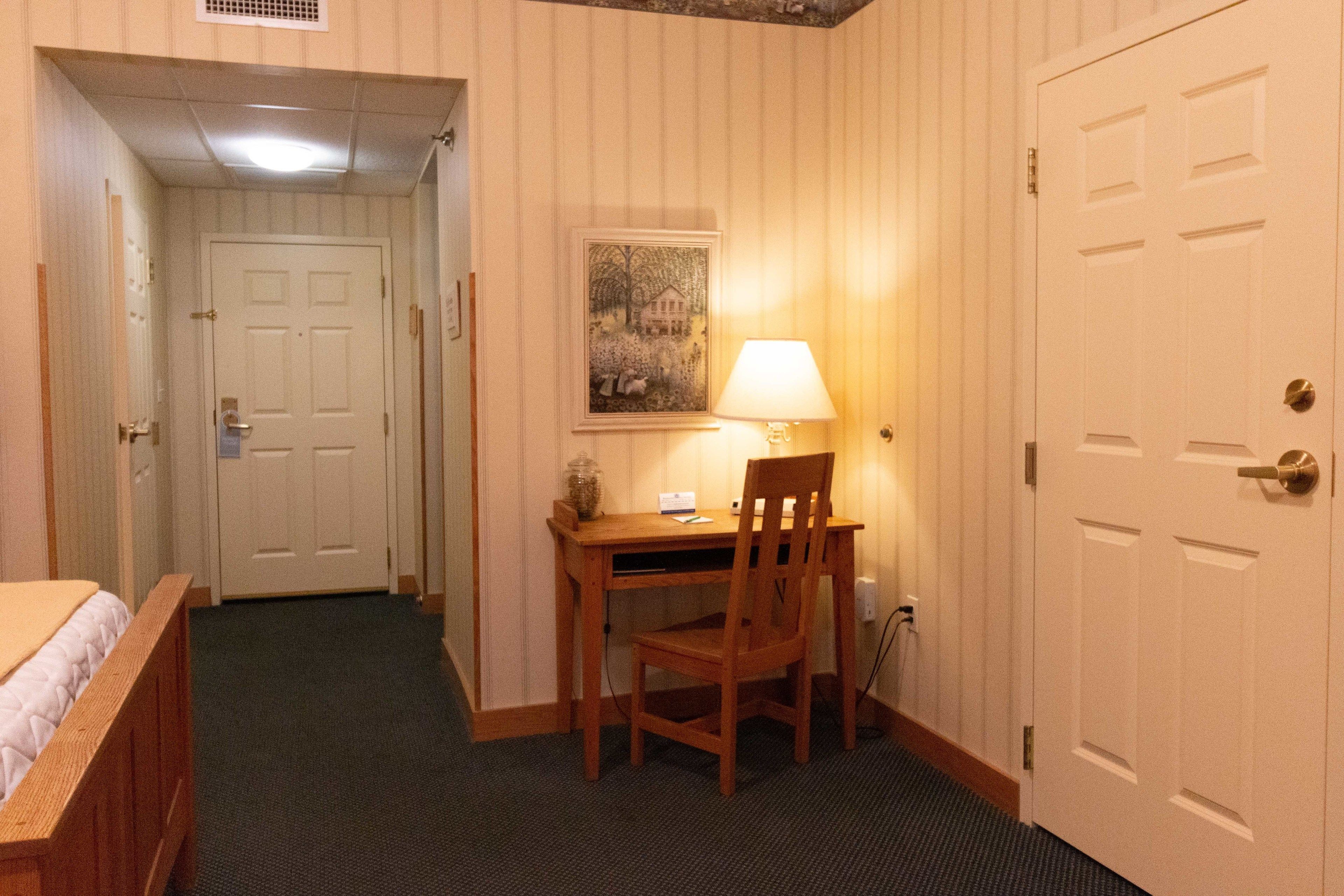 A hotel room with a bed, a desk with a chair, a lamp, and a door.