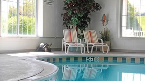 An indoor swimming pool with two striped lounge chairs, a potted plant, and a sign indicating pool capacity.