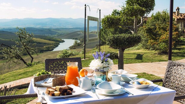 Une table de jardin dressée pour un repas en plein air, avec de la nourriture et des boissons, offrant une vue panoramique sur un paysage pittoresque, avec une rivière et des collines.