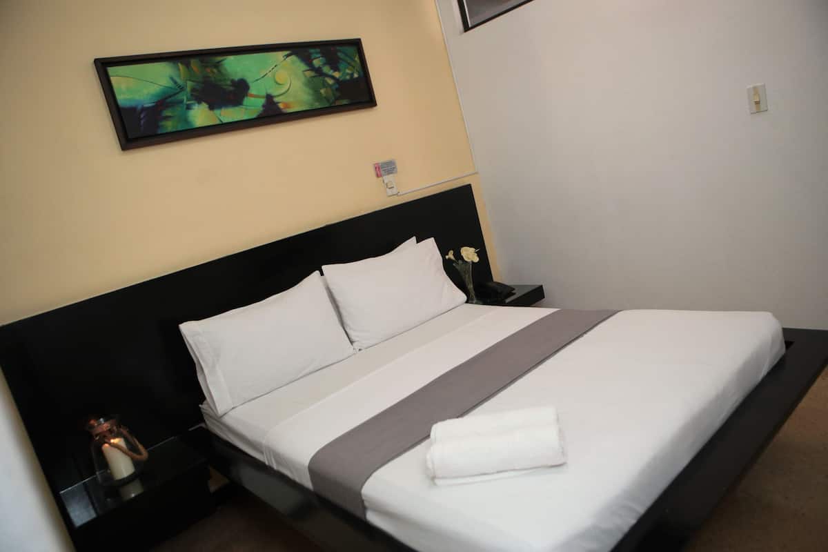 A neatly made bed with white linens and a gray accent, a nightstand with a candle, and a framed abstract painting on the wall.