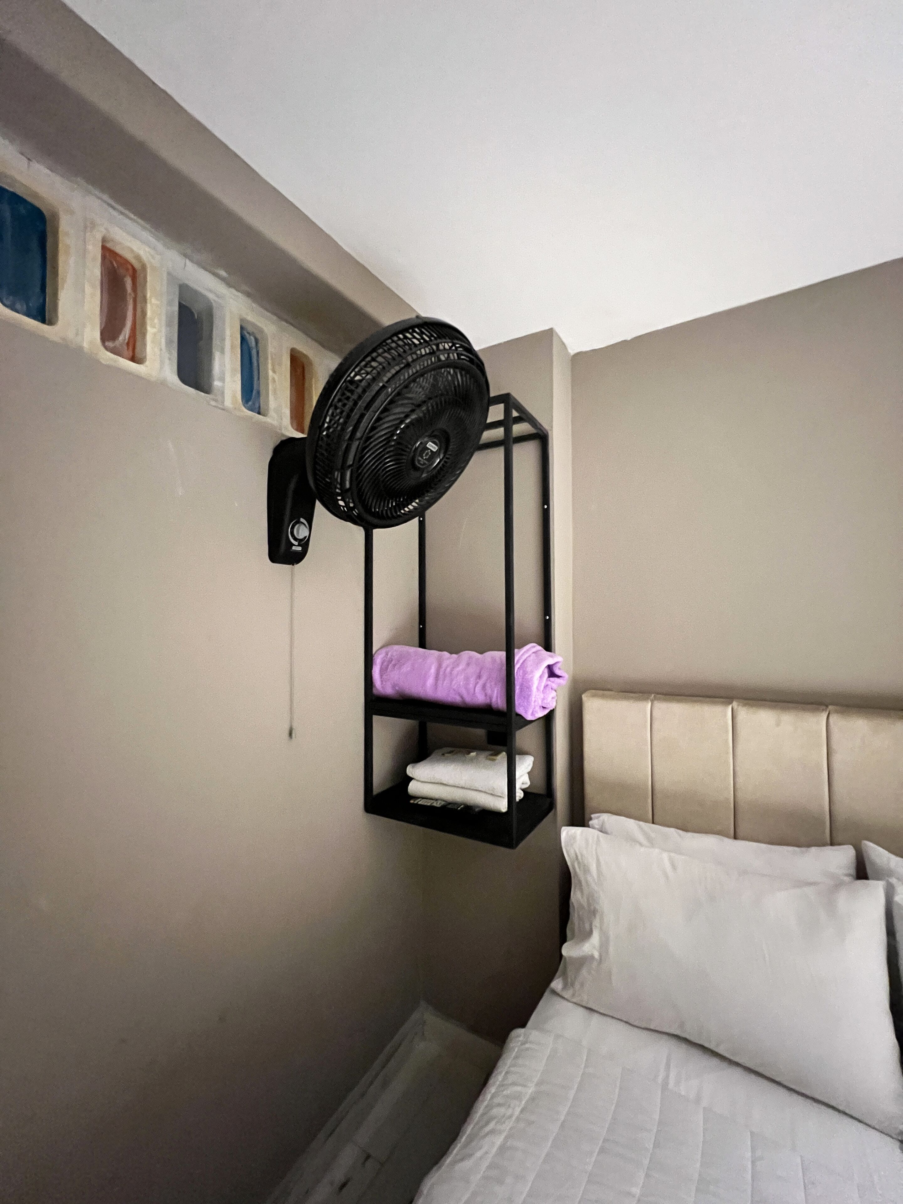 Une petite pièce comprenant un lit, un ventilateur fixé au mur et une étagère avec des serviettes pliées.
