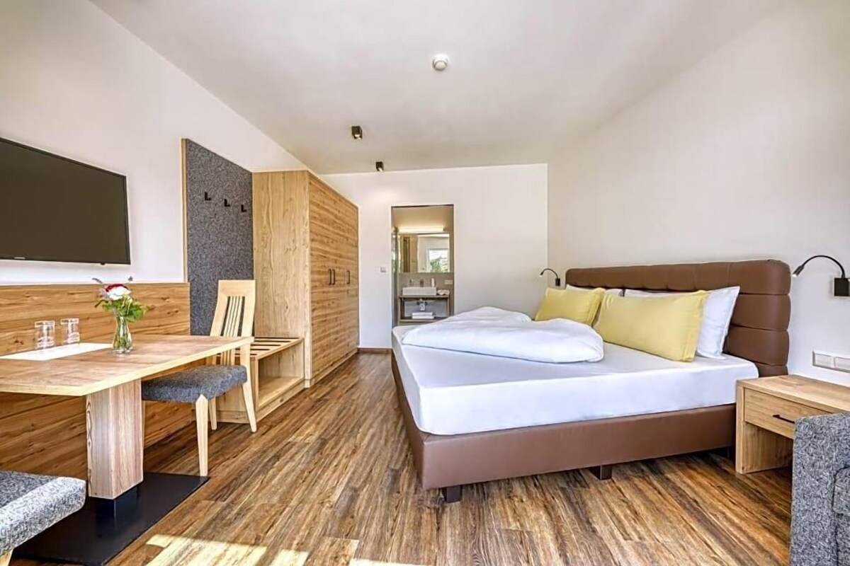 Habitació d'hotel moderna amb sòl de fusta, un llit amb roba de llit blanca, una taula de treball amb una cadira i un televisor de pantalla plana penjat a la paret.