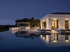 A modern outdoor pool area with lounge chairs and a seating area, surrounded by a well-maintained garden and a building with large windows. - Drallos Luxury Villa (Zakynthos)
