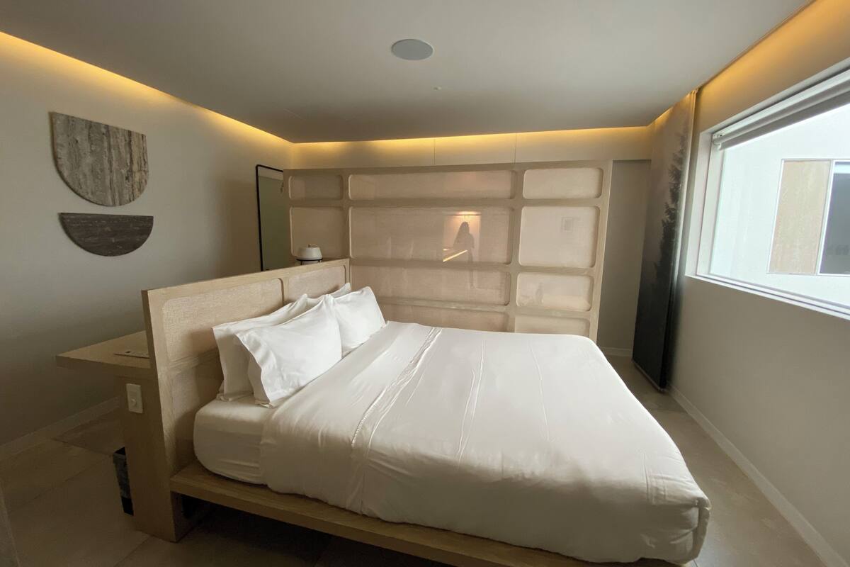 A modern bedroom with a large bed, a nightstand, a wall-mounted mirror, and a window providing natural light.