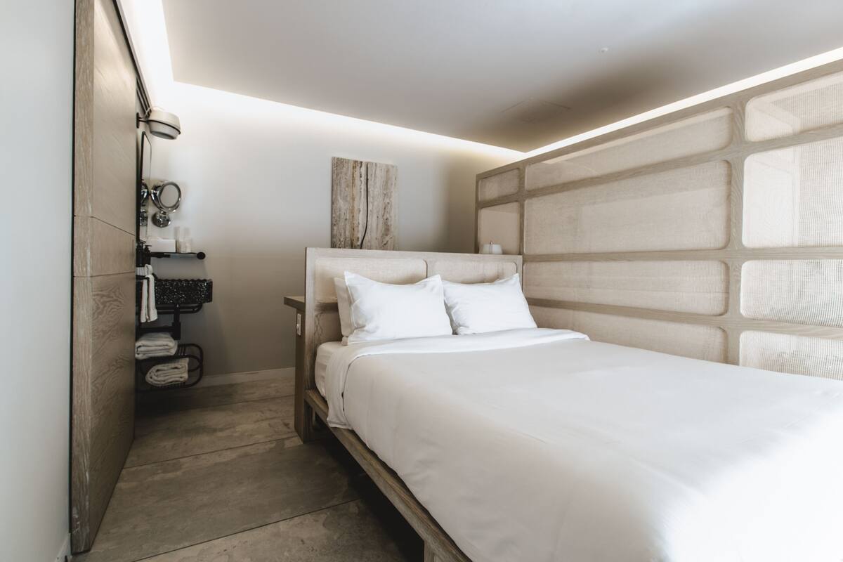 A modern bedroom with a large bed, a headboard with a geometric pattern, a bedside table with a mirror, and a towel rack.