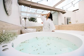 Persoon in een witte badjas bij een kuub met helder water, omringd door een modern interieur met een pergola en potplanten.