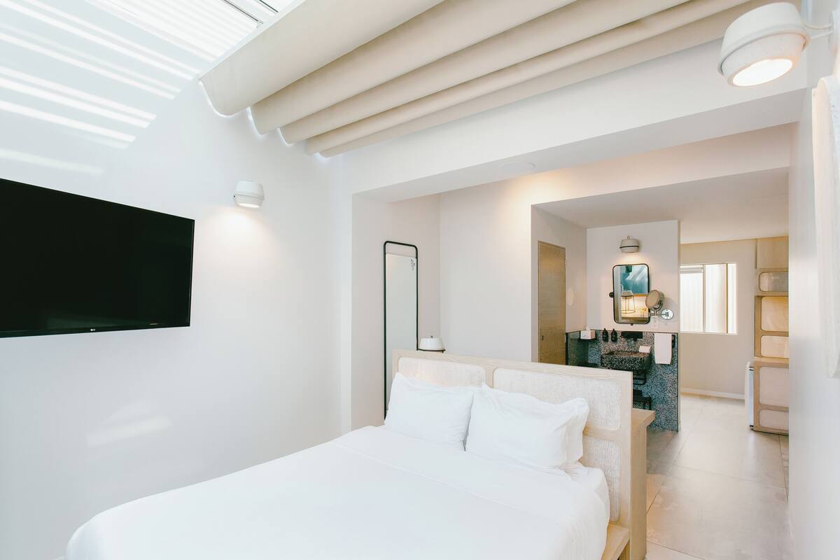 A modern hotel room with a large bed, a flat-screen TV mounted on the wall, a walk-in closet, and a bathroom with a mirror and sink.