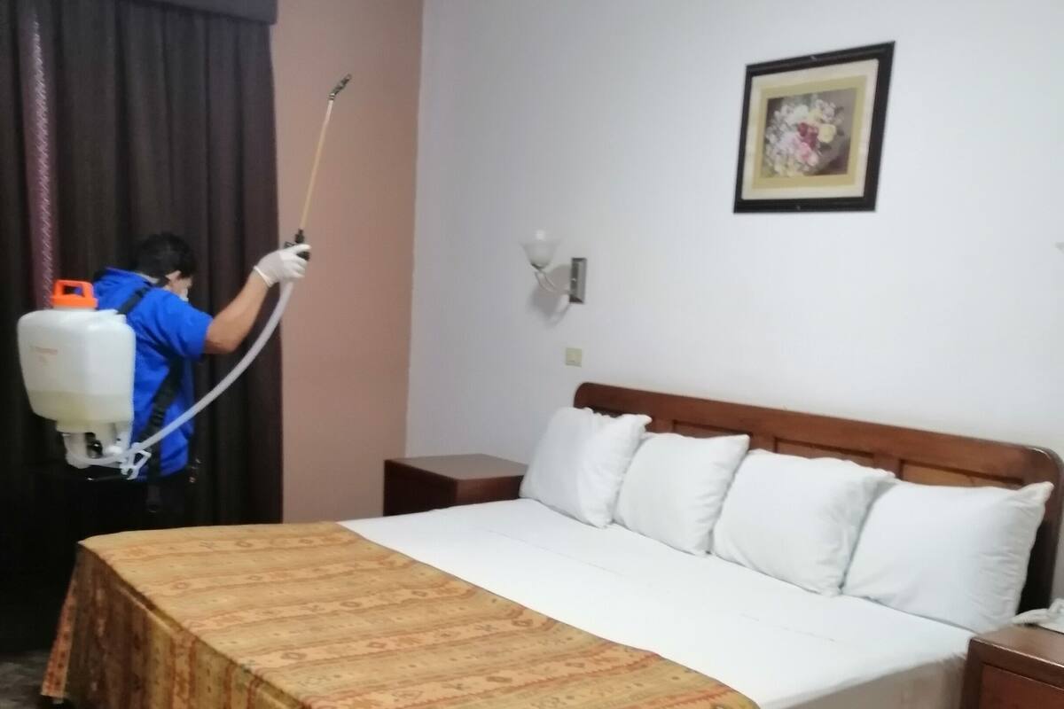 Habitación de hotel con cama, mesita de noche, lámpara de pared y una persona limpiando el suelo con un pulverizador.