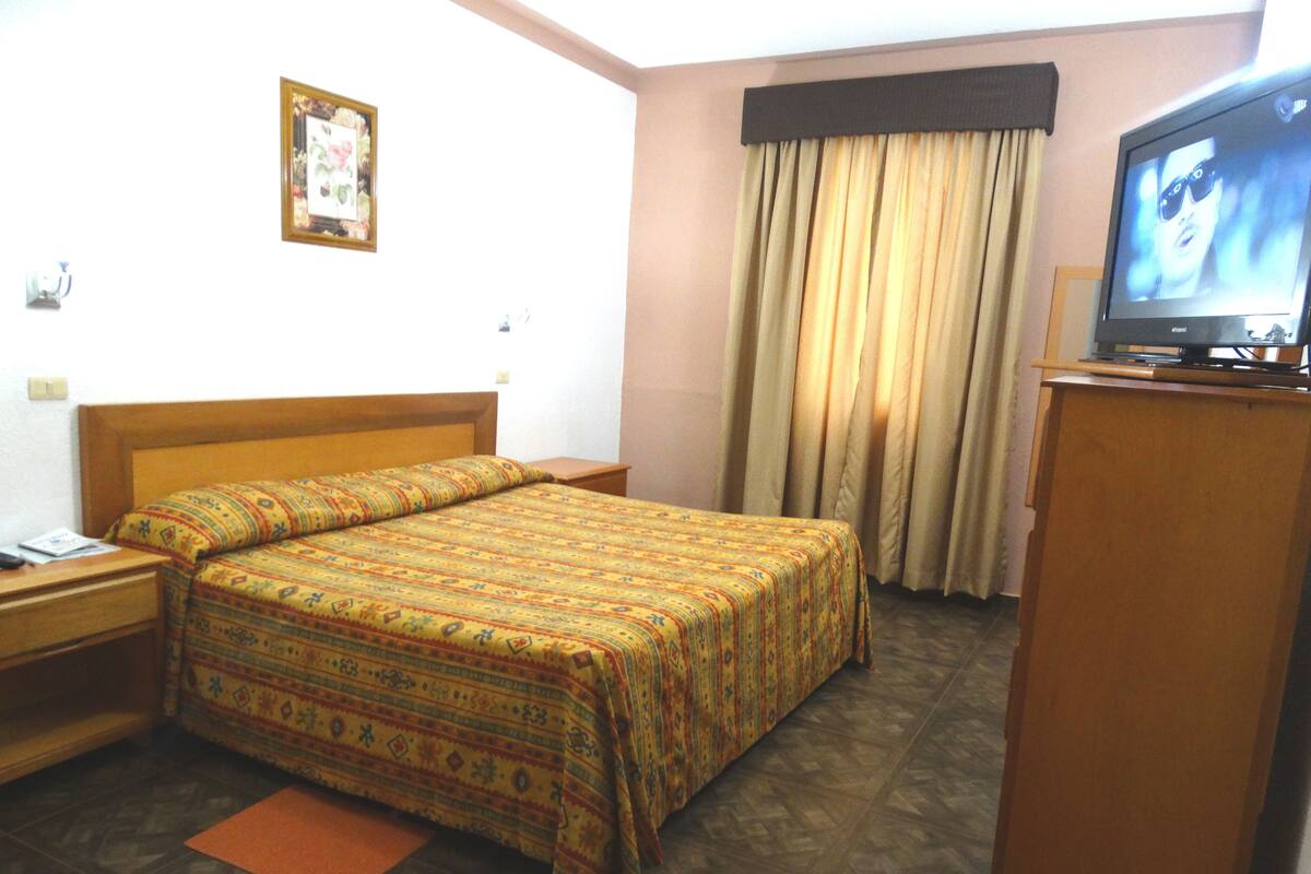 Una habitación de hotel con cama, televisión, escritorio y ventana con cortinas.