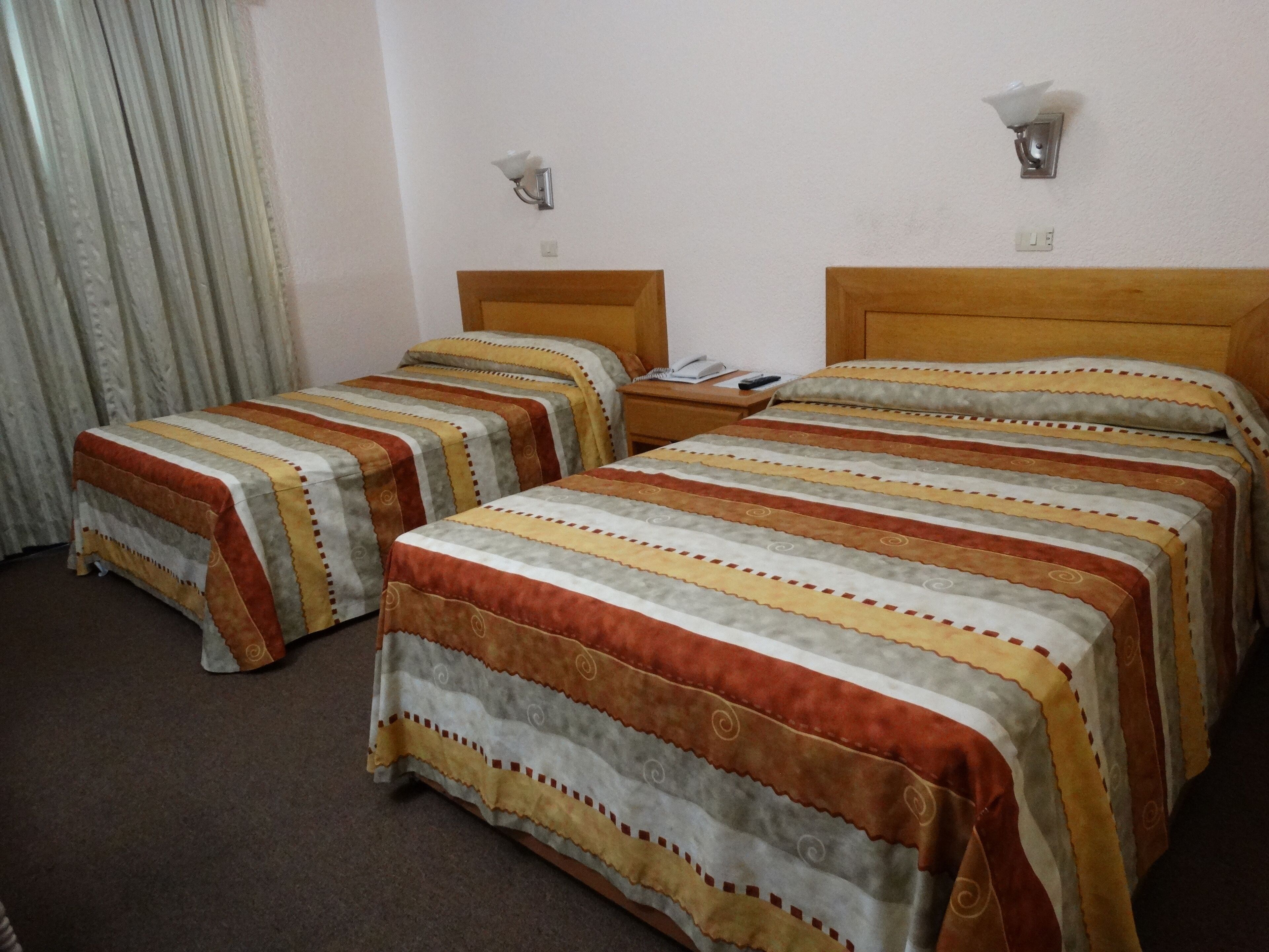 Habitación de hotel con dos camas, cada una con ropa de cama a rayas, cabecero de madera y mesita de noche con lámpara.