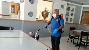 A person cleaning a table with a spray bottle in a room with tiled floors, a large window, and decorative plates on the wall. - Hotel Astromundo (Reynosa)