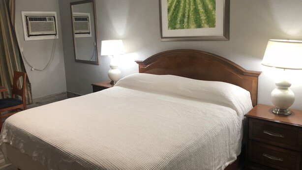 A hotel room with a neatly made bed, a wooden headboard, a nightstand with a lamp, a wall-mounted air conditioner, a mirror, and a framed picture above the bed.