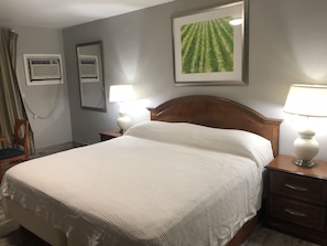 A hotel room with a neatly made bed, a wooden headboard, a nightstand with a lamp, a wall-mounted air conditioner, a mirror, and a framed picture above the bed. - Bay Breeze Inn Bradenton Downtown (Bradenton)