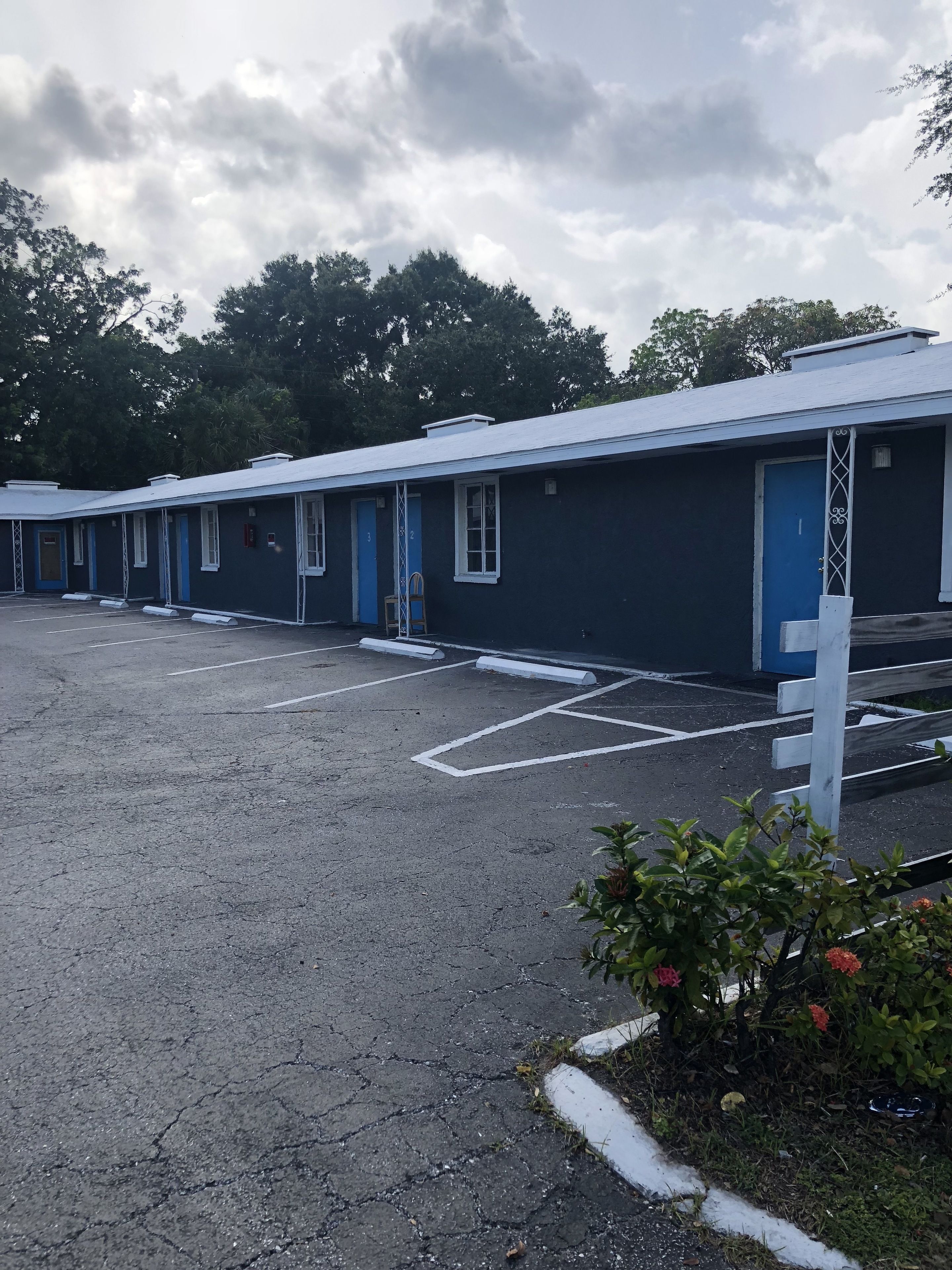 A row of motel rooms with blue doors and white trim, a parking lot, and a garden area with flowers.