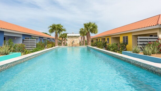 A rectangular swimming pool with clear blue water, surrounded by buildings with red-tiled roofs and a stone wall in the background. - Centro Garden (Willemstad)