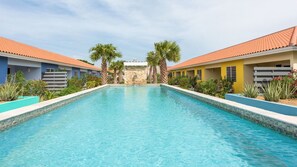 A rectangular swimming pool with clear blue water, surrounded by buildings with red-tiled roofs and a stone wall in the background. - Centro Garden (Willemstad)