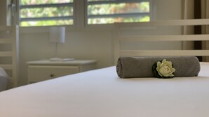 A neatly made bed with a rolled towel and a flower, a white nightstand, and a window with blinds. - Centro Garden (Willemstad)