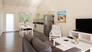 A modern living room with a grey sofa, a dining table with chairs, a kitchen area with a fridge, and a TV on a stand. - Centro Garden (Willemstad)
