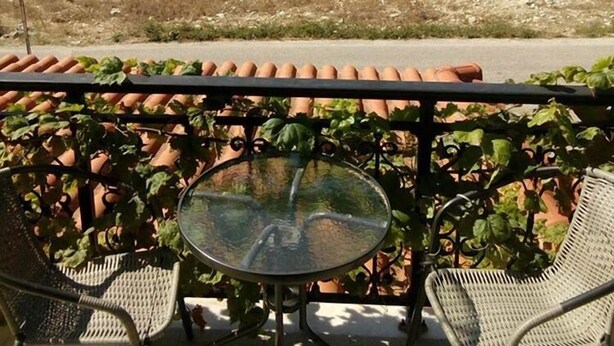 Ein Balkon mit Glastisch und zwei Korbstühlen, der auf eine hügelige Landschaft mit Straße und Vegetation blickt.
