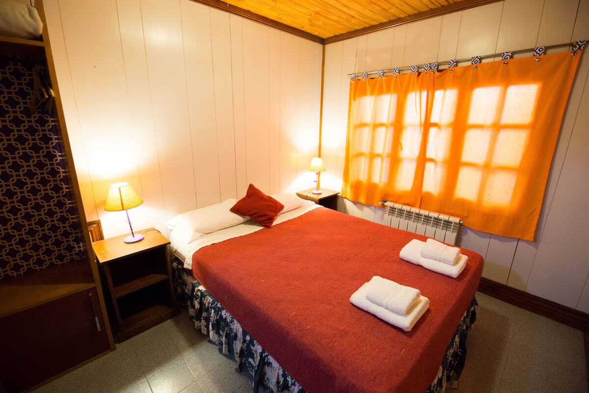 A hotel room with a bed, two bedside lamps, a window with curtains, a radiator, and a patterned wall.
